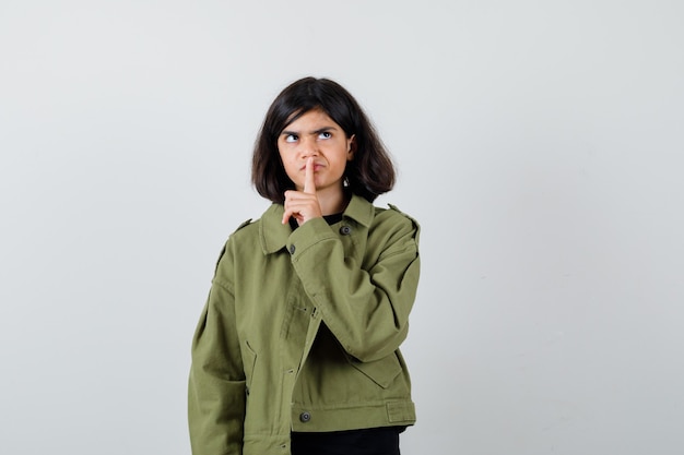 Teen girl showing silence gesture in t-shirt, green jacket and looking focused. front view.