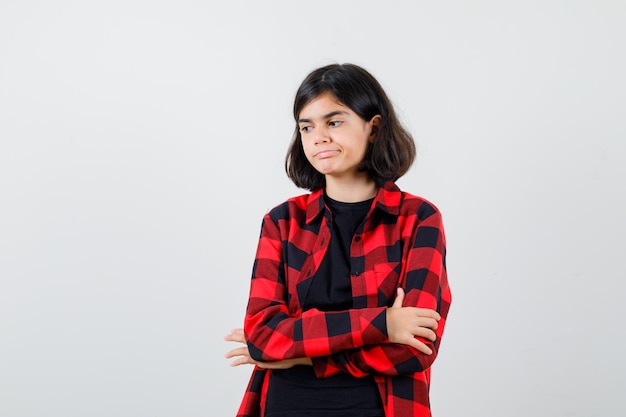 Teen girl in t-shirt, checkered shirt standing with crossed arms and looking pensive , front view.