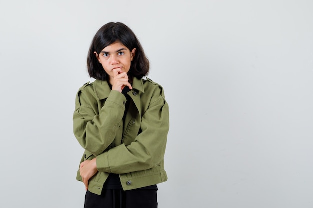 Teen girl in t-shirt, green jacket propping chin on hand and looking careful , front view.