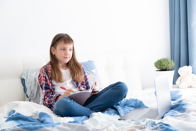 teen girl watching videos on laptop sitting on bed in her room. teenage school student studying online, distance learning,