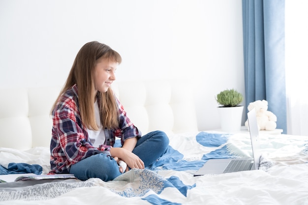 teen girl watching videos on laptop sitting on bed in her room. teenage school student studying online, distance learning,