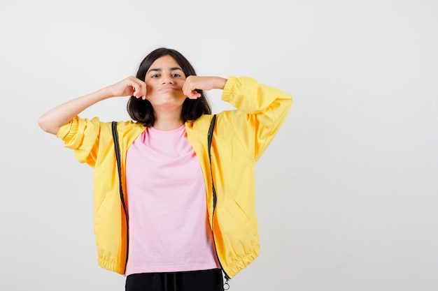 Teen girl in yellow tracksuit, t-shirt pulling cheeks to the sides and looking wistful , front view.