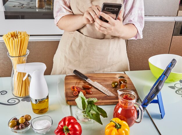 Photo teen prepares virtual online textbook tutorial and watches digital recipe on touch screen mobile phone while preparing healthy food in kitchen at home