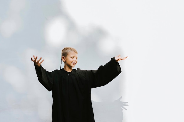 teenage boy in clothes of a graduate coat and cap celebrates high school or junior year graduation on background of white wall with shadows and with diploma in hands education and no school concept