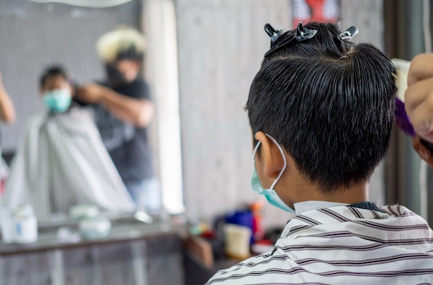 Teenage boy in a face protective mask is getting a haircut from a barbershop .