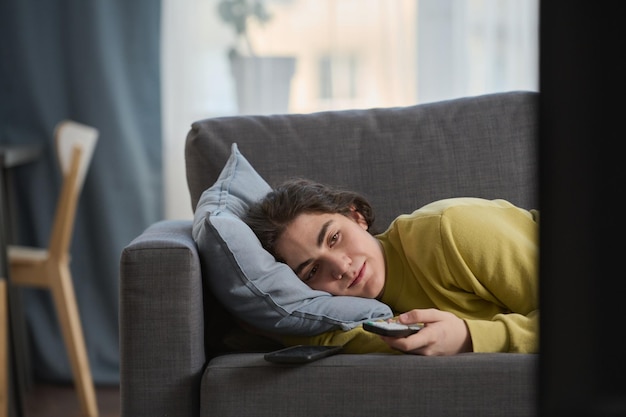 Teenage boy lying on sofa and switching channels with remote control while watching tv in the room