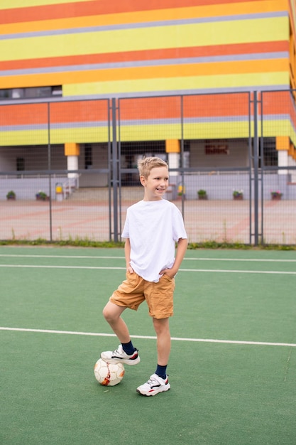 A teenage boy stands on a green field in the school yard with a soccer ball and looks away