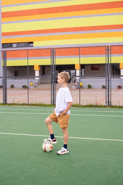 A teenage boy stands on a green field in the school yard with a soccer ball