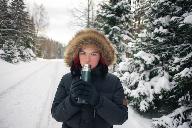 Teenage boy walks through a snowy forest on a cold winter day with a hot drink in a thermos. Healthy lifestyle. Useful activity.