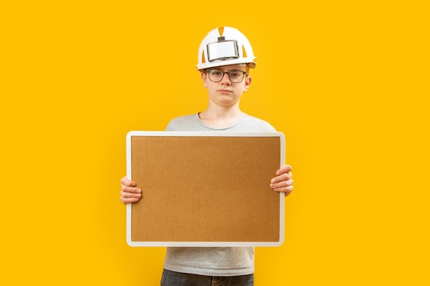 Teenage boy wears glasses and protective helmet holding blank cork board on yellow background Copy space Mock up