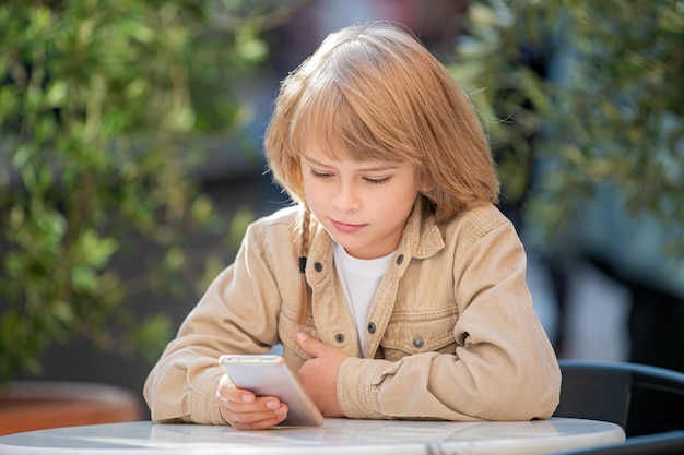 Teenage boy with a smartphone in his hands sits at a table in summer cafe