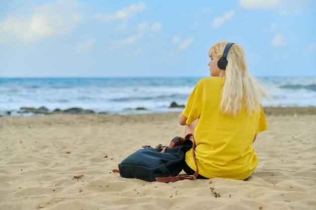 Teenage female in headphones with backpack sitting on beach back view copy space