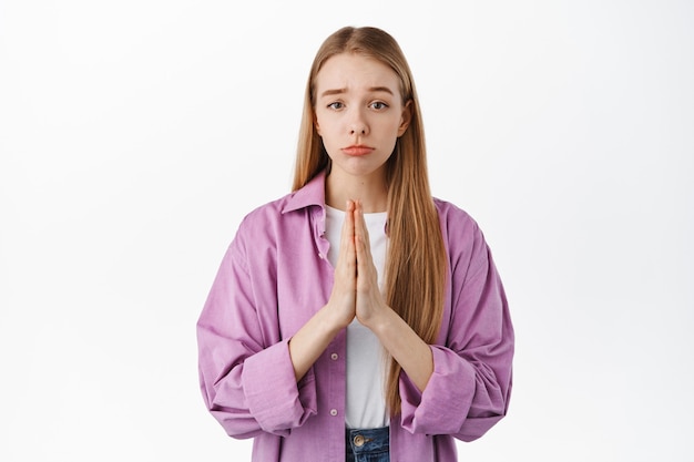 Teenage girl asking for favour, begging, say please with hands in pray beg gesture, frowning and looking sad, standing against white wall