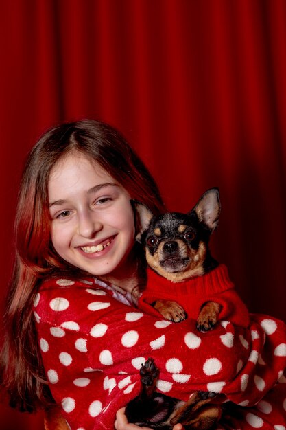 Teenage girl and a chihuahua dressed in a red sweater for dogs at home against a background of red.