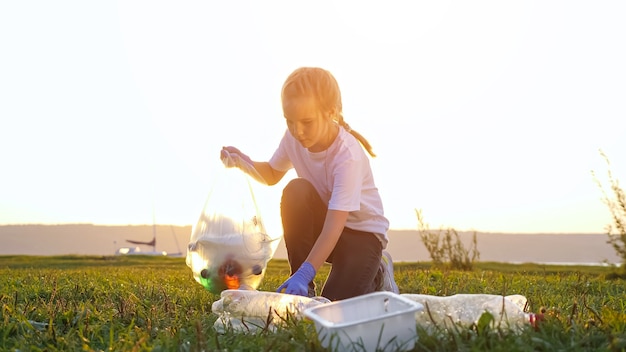 Teenage girl collects rubbish in gloves on the river bank at sunset.