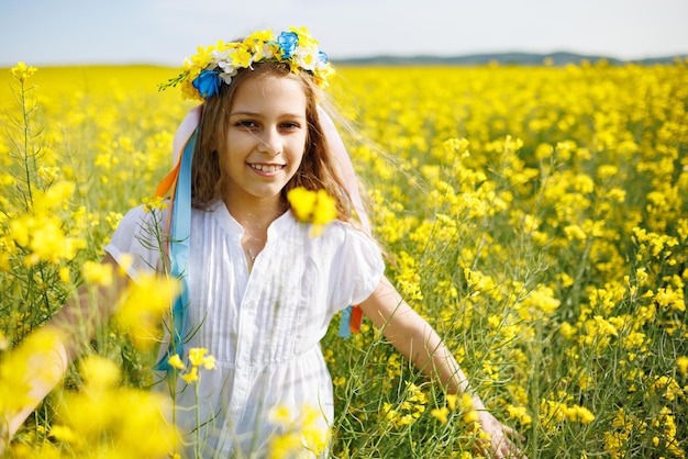 Teenage girl in dress and Ukrainian wreath with lentims on head in rapeseed field under a clear blue sky