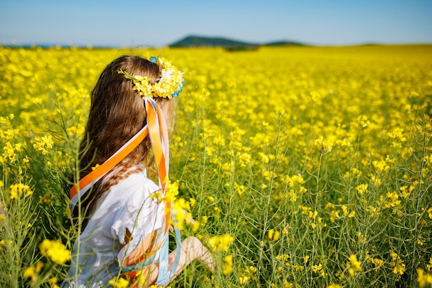 Teenage girl in dress and Ukrainian wreath with lentims on head in rapeseed field under a clear blue sky