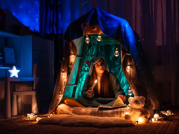 Teenage girl in the form of anime sitting in the game home tent. Scenery with fantastic garland lighting 