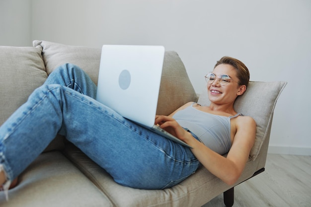 Teenage girl freelancer with laptop sitting on couch at home smiling in home clothes and glasses with short haircut lifestyle with no filters free copy space