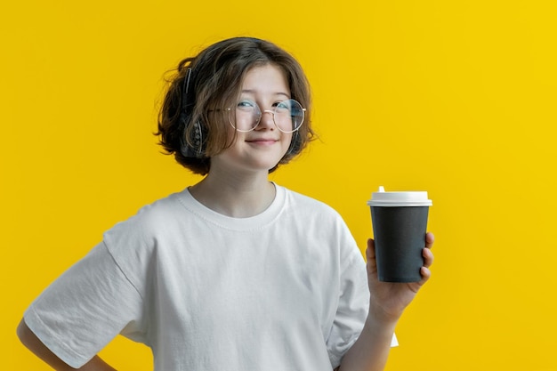 Teenage girl in glasses headphones with cup of coffee on yellow background