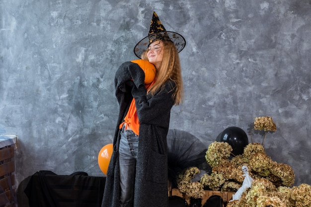 Photo teenage girl in hat among decor for halloween holiday, huge pumpkin in hand. humor photo