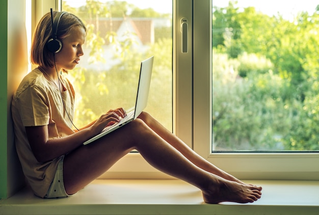 Teenage girl in headphones using laptop and sitting on the window