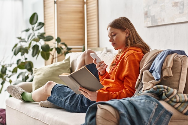 teenage girl holding book and using smartphone sitting on sofa next to messy pile of clothes