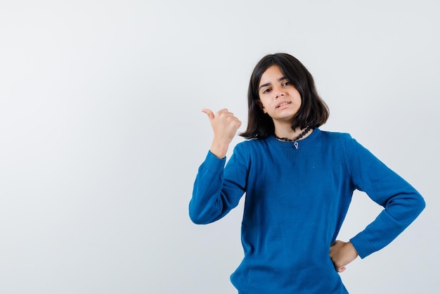 The teenage girl is pointing to left with thumb and putting other hand on waist on white background