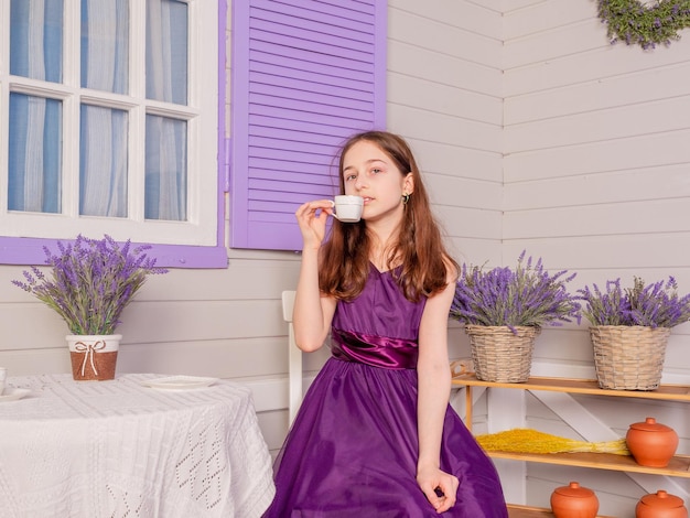 A teenage girl in a purple dress drinks tea from a white cup. The girl is sitting at the table.