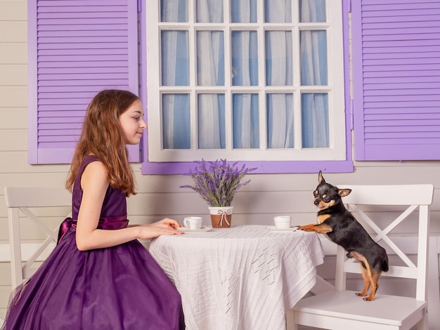 A teenage girl in a purple dress drinks tea from a white cup with a chihuahua dog.