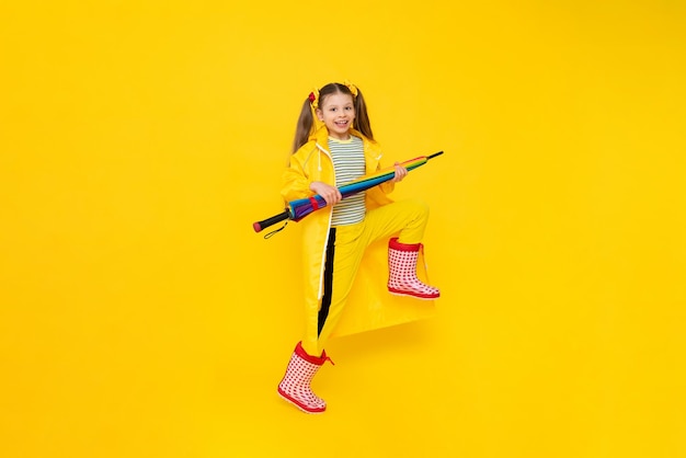 A teenage girl in a raincoat and rubber boots walks on a yellow isolated background Spring autumn rainy weather Full size profile side photo of a happy walking little girl