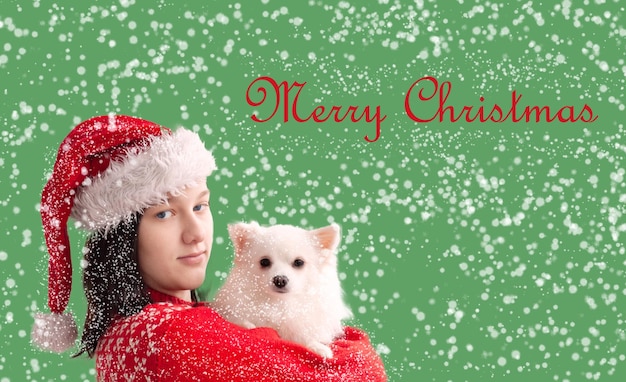 A teenage girl in a Santa Claus hat and a red Christmas sweater holds a white pomeranian in her arms The concept of Christmas and New Year