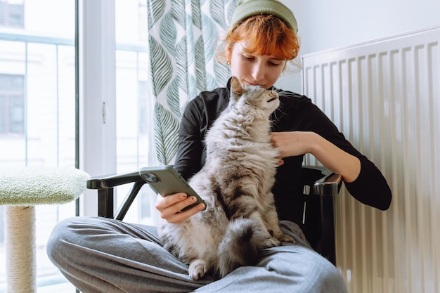 Teenage girl sitting on chair holding cat and looking at phone