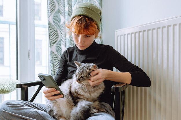 Teenage girl sitting on chair holding cat and looking at phone