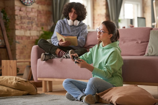 Teenage girl sitting on the floor and playing video game with boy reading a book on sofa in the background