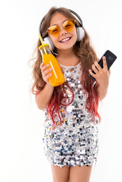 Teenage girl in sparkling dress and yellow sunglasses, with big white earphones with black smartphone and bottle with orange juice in her hands isolated