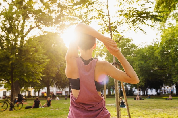 teenage girl in sports shorts and top does back stretch in public park in sun. Healthy active