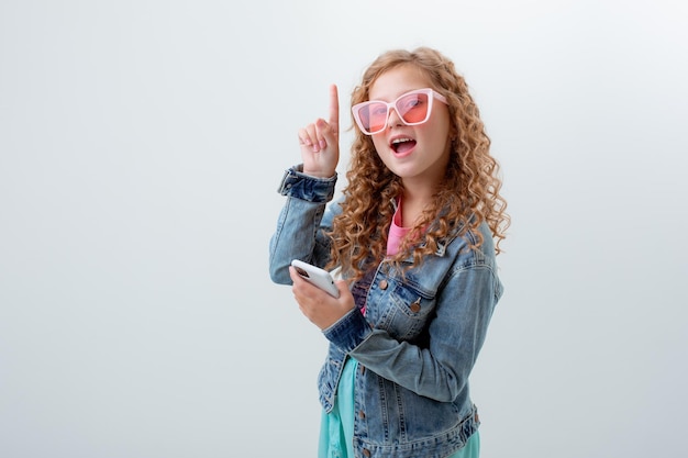 A teenage girl in sunglasses holding a phone shows surprise on a white background