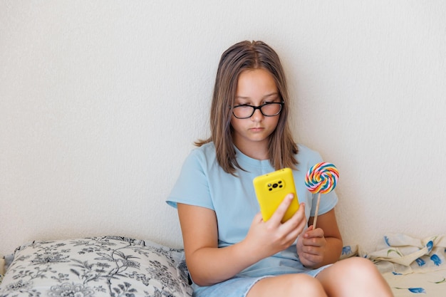 Teenage girl talks on the phone with a lollipop in her hand sitting on the bed