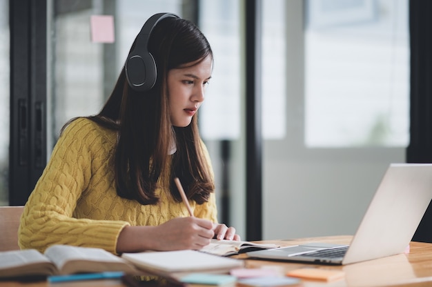 Teenage girl wearing headphones studying online from home, She looks at a laptop and taking notes, studying online.