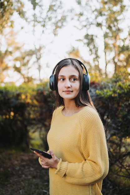 Teenage girl wearing yellow sweater, using headphones for playing music on her smartphone outdoor.