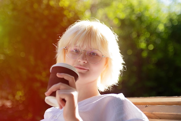 Teenage girl with blond hear drinking coffee from craft paper cup outsidesunlight on background