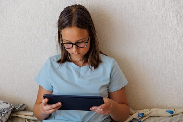 Teenage girl with glasses playing a tablet on a bed in the bedroom