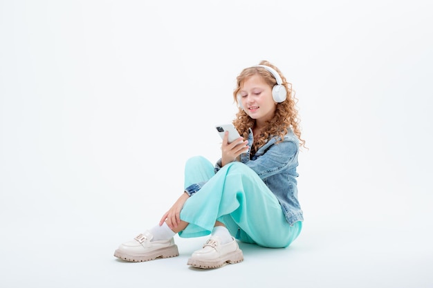 A teenage girl with headphones holding a phone sits on a white background