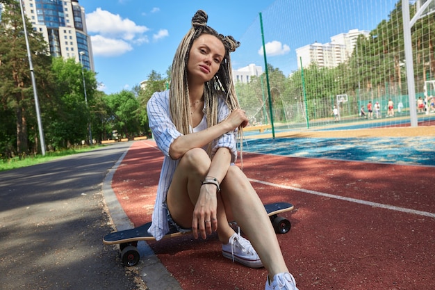 A teenage girl with long pigtails sits on a longboard near a sports ground around green trees high q...