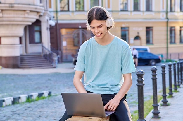 Teenage male student in headphones using laptop outdoor