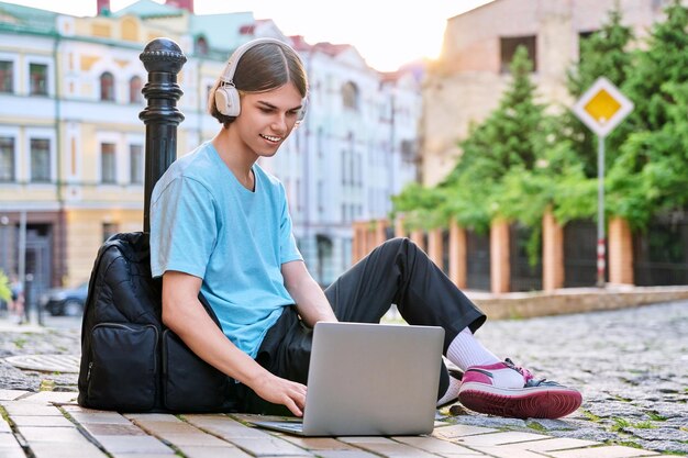 Teenage male student in headphones using laptop outdoor