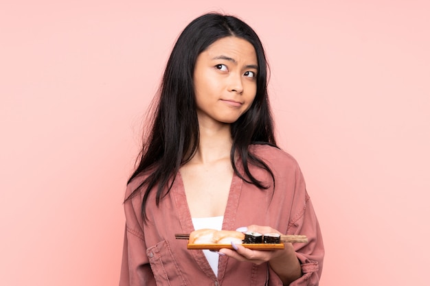 Teenager Asian girl eating sushi on pink wall standing and looking to the side