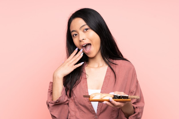 Teenager Asian girl eating sushi on pink wall with surprised facial expression