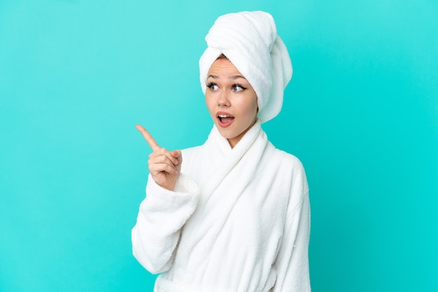 Teenager blonde girl in a bathrobe over isolated blue background intending to realizes the solution while lifting a finger up